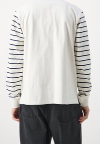 Dlouhý rukáv, bílá košile se námořnickými horizontálními pruhy na rukávech, vyrobená z bavlny. Má volný střih a žebrované manžety.