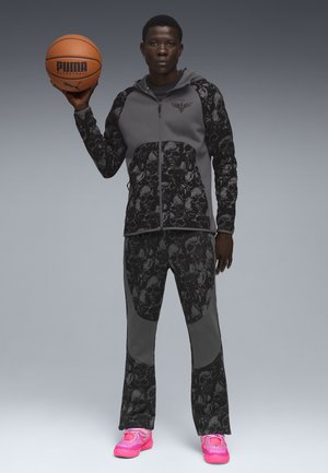 Basketball outfit bestående af en grå hættetrøje med lynlås og dødningehovede accentuerende detaljer samt matchende bukser, parret med lyserøde sneakers.