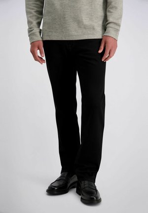 Man draagt zwarte rechte broek, zwarte leren loafers en een lichtgrijs hemd met lange mouwen, staand tegen een effen achtergrond.