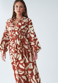 Maksi haljina s hrđavo-narančastom pozadinom i kremastim apstraktnim uzorcima. Ima prednji dio na kopčanje s gumbima, remen oko struka i voluminozne rukave.