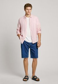 Svetlo roza lanena srajca s gumbi preko bele majice, v kombinaciji z jeans kratkimi hlačami in temnimi sandali. Model ima kodraste lase.