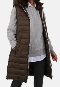 Woman wearing a long dark brown quilted sleeveless coat over a grey sweatshirt, white shirt, and black pants, standing with hands relaxed.