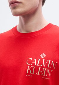 Κόκκινο βαμβακερό T-shirt με στρογγυλή λαιμόκοψη. Η φράση "CALVIN KLEIN" τυπωμένη σε χρυσό, με τις λέξεις "W. 39TH ST." και "NEW YORK CITY" από κάτω σε μικρότερο κείμενο.