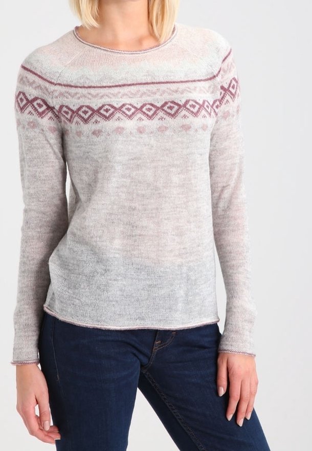 Femme portant un pull en maille gris clair avec un motif géométrique bordeaux sur la poitrine et un jean bleu foncé, debout devant un fond uni.