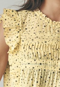Żółta bluzka z tkaniny w czarne kropki, z falbaniastymi rękawami i marszczonymi detalami na dekolcie.