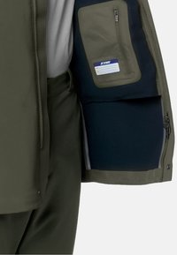 Giacca impermeabile verde con tasca esterna con zip e tasca interna foderata in pile. Presenta un'esterno liscio e un morbido tessuto interno.