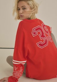 Červená oversized mikina s bílými pruhy na rukávech, zdobená dekorativními květinovými výšivkami a velkým číslem "39" na zádech.
