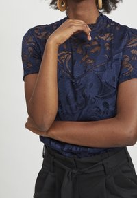 Marinblus med korta ärmar och blommönster, texturerat tyg, hög hals, matchad med svarta höga byxor.