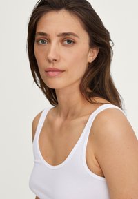Witte tanktop met dunne schouderbanden en een ronde halslijn. De stof lijkt soepel en aansluitend, wat de schouders accentueert.