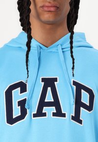 Light blue hoodie featuring a large navy "GAP" logo with white outline. Drawstrings with metal tips. Soft, textured fabric.