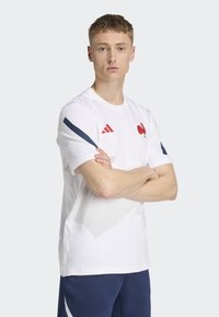 Camiseta blanca de manga corta con acentos rojos. Presenta un logo de gallo rojo y rayas azules en los hombros. Tejido suave, escote redondo.