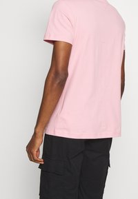 Camiseta de algodón rosa claro con escote redondo y mangas cortas. La parte posterior presenta un pequeño detalle con el logo y costuras limpias a lo largo del dobladillo.