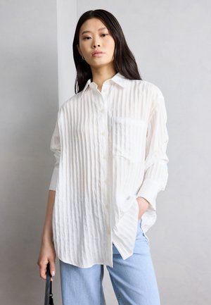 Młoda kobieta o długich ciemnych włosach, ubrana w luźną, białą, teksturowaną koszulę na guziki i jasnoniebieskie dżinsy, trzymająca czarną torebkę.