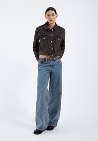 Donna in piedi che indossa una giacca di jeans nera corta, jeans larghi blu con cintura borchiata e scarpe nere su uno sfondo chiaro uniforme.
