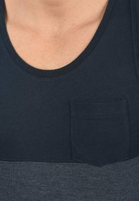 Tamnoplava majica bez rukava sa okruglim izrezom, kontrastnim sivim donjim dijelom i malim džepom na grudima s teksturiranim završetkom.