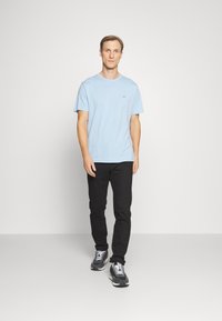 Hellblaue Baumwoll-T-Shirt mit rundem Halsausschnitt, kombiniert mit schwarzen Hosen und grauen Sneakers, die weiße Schnürsenkel und ein modernes Design aufweisen.