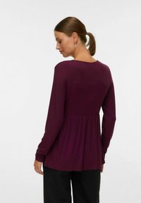 Haut à manches longues en velours prune avec un col rond et une taille plissée. Le tissu a une texture douce et la silhouette est décontractée.
