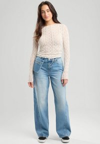 Witte kanten top met lange mouwen en een aansluitende taille, gecombineerd met lichtblauwe wijde jeans met een zijzak.