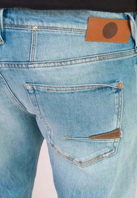 Blauwe denim jeans met een achterzak met een bruin leren label, een versleten textuur en contrasterende stikseldetails.