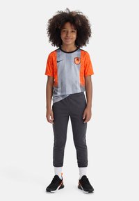 Παιδί με σγουρά μαλλιά στέκεται φορώντας γκρι και πορτοκαλί αθλητική μπλούζα, σκούρα φόρμα, λευκές κάλτσες και μαύρα αθλητικά παπούτσια σε λευκό φόντο.