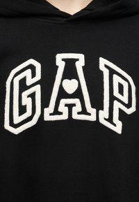 Μαύρο ένδυμα με μεγάλα λευκά ανάγλυφα γράμματα "GAP", με το γράμμα "A" να έχει ένα μικρό καρδιά στο κέντρο.