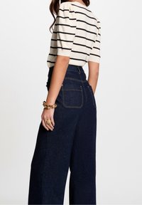 Creme- og sort stribet top med puffærmer, parret med højtraljede mørk denim bukser med vide ben, der har baglommer og guld accessories.