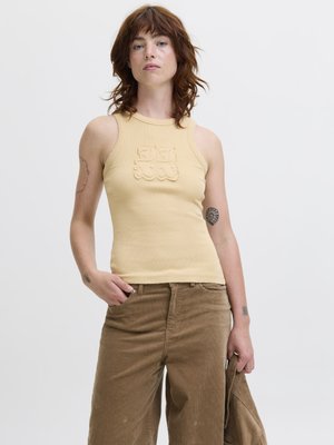 Femme aux cheveux roux portant un haut beige sans manches et un pantalon marron, tenant une veste marron assortie sur un fond blanc uni.