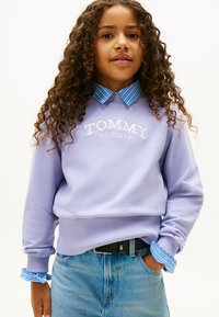 Liliowa bluza z prążkowanym wykończeniem i haftowanym logo "TOMMY HILFIGER", noszona na niebieskiej i białej w paski koszuli z kołnierzykiem. Dżinsy i czarny pasek.