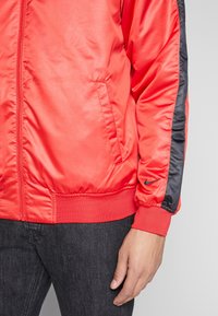 Czerwona kurtka na zamek z czarnym paskiem na rękawie i logo Nike przy nadgarstku, noszona z ciemnymi dżinsami, krótka do talii, z widoczną dłonią.