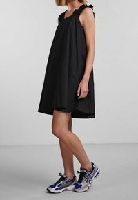 Svart ärmlös klänning med volangdetaljer på axelbanden, lös A-linjeformad silhuett, kombinerad med blå och silverfärgade sportskor.