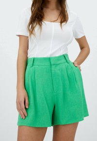 Pantaloni corti in lino verde con pieghe, vita alta e tasche laterali, abbinati a una maglietta bianca aderente con maniche corte e scollo quadrato.