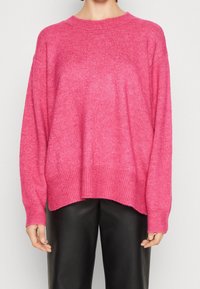 Osoba w luźnym różowym swetrze dzianinowym i czarnych skórzanych spodniach, widoczna od ramion do bioder na jednolitym tle.