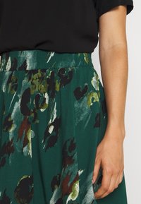 Grüne Rock mit floralem Muster, das schwarze, dunkelgrüne und metallische Akzente aufweist. Elastischer Bund und fließender Stofftextur.