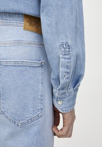 Camisa de mezclilla azul claro con mangas largas, puños abotonados y una etiqueta de cuero en la parte posterior. Presenta costuras sutiles y un corte clásico.