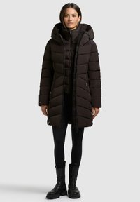 Giacca a vento nera con cappuccio, con sezioni trapuntate, cerniera frontale e regolatori a cordoncino. Indossata sopra leggings neri e stivali neri.