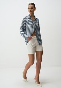 Gestreepte blouse in blauw en wit, gecombineerd met witte shorts en beige hakken. Soepele stof, losse pasvorm en lange mouwen.