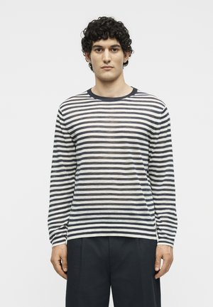 Άνδρας με κοντά σγουρά μαλλιά που φοράει μαύρο και άσπρο οριζόντια ριγέ μπλουζάκι με μακριά μανίκια και σκούρο παντελόνι, στέκεται αντικρίζοντας μπροστά.