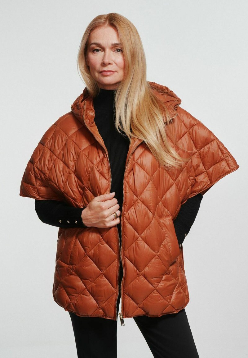 Brązowa puchowa kurtka z kapturem, pikowana, z zapięciem na zamek z przodu, szerokimi rękawami i gładkim połyskiem. Nosi się na czarnym golfie.