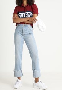 Polo a righe rosse e blu con colletto classico. Jeans azzurri a vita alta con orli sfrangiati. Sneakers bianche e cappellino bianco.