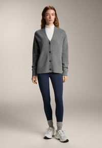 Szürke kötött kardigán V-nyakkivágással, négy fekete gombbal. Sötétkék leggingsszel és fehér tornacipővel párosítva. Simás tapintású.