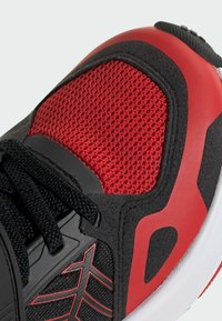 Crne i crvene atletske tenisice s teksturiranim mrežastim gornjim dijelom, crnim vezicama, glatkim crvenim detaljem na peti i bijelim potplatom s utorima za bolje prijanjanje.