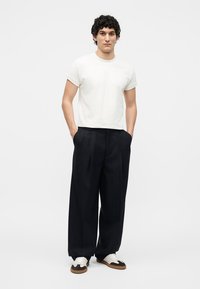Man wearing a plain white short-sleeve shirt, loose black pleated trousers, and white and black sneakers, standing with hands in pockets.