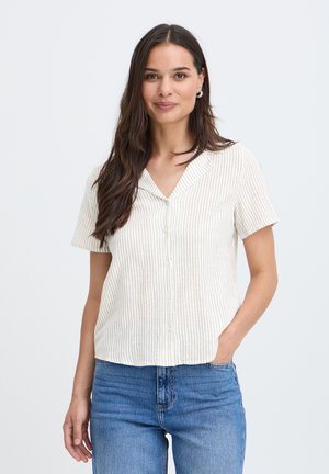 Kvinde med langt brunt hår bærer en kortærmet, cremefarvet stribet bluse og blå jeans, står med den ene hånd i lommen og smiler let.