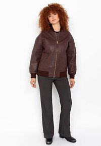 Veste aviateur en cuir marron avec une fermeture éclair frontale, des poignets et un ourlet côtel és, et une poche zippée sur la manche gauche. Texture lisse.