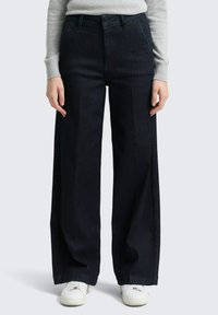 Pantalon large en denim foncé taille haute avec plis avant, deux poches latérales, et une finition soignée. Associé à des baskets blanches.