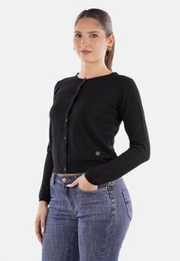 Cardigan lungo nero con maniche lunghe, colletto rotondo e bottoni frontali. Indossato sopra jeans blu, con un sottile accento del logo.