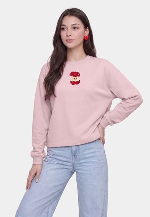 Ung kvinde med langt mørkt hår iført en lyserød sweatshirt med et rødt æblekernehave-design og lyseblå jeans, står med den ene hånd på hoften.