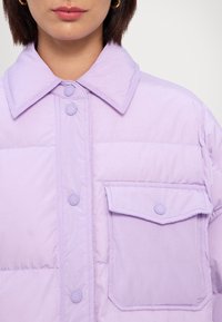 Chaqueta acolchada lavanda con cierre de botones en la parte delantera, gran cuello y un bolsillo en el lado izquierdo del pecho, con botones mate y textura acolchada.