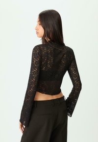Top corto in pizzo nero con maniche lunghe, bordi scallop e design trasparente, abbinato a pantaloni neri a vita alta.