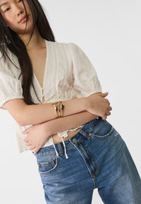 Witte blouse met pofmouwen, V-hals en knoopdetails. Donkerblauwe spijkerbroeken met een lichte vervaging. Gouden armband met golvend ontwerp.